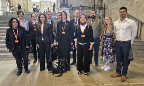 A group of around twelve people wearing name badges stand together inside a large, historic hall with stone walls, tall windows and steps behind them,