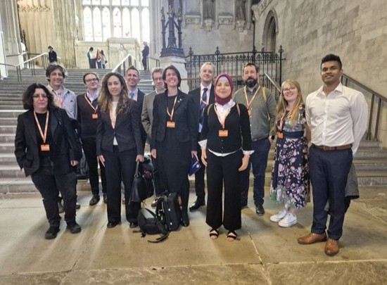 A group of around twelve people wearing name badges stand together inside a large, historic hall with stone walls, tall windows and steps behind them,