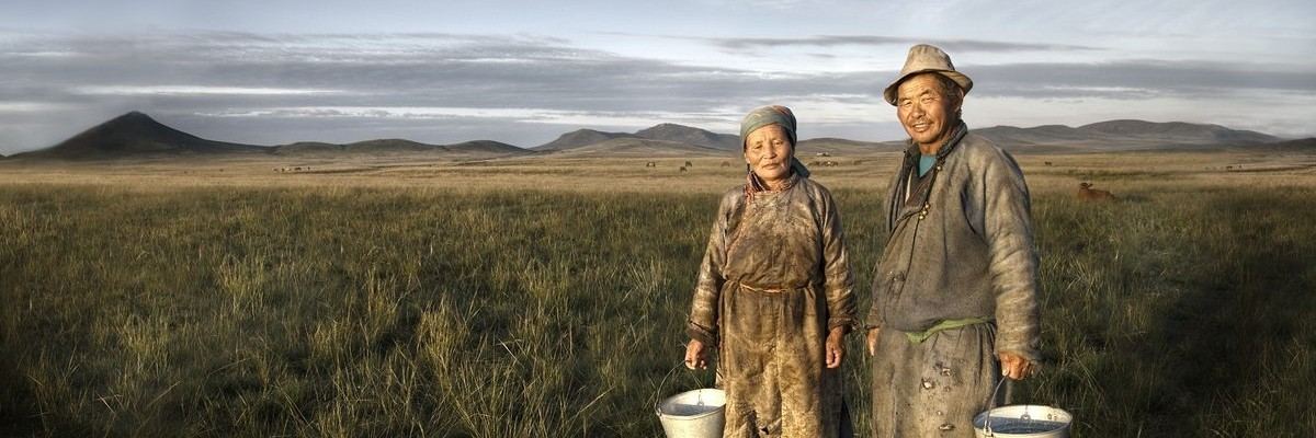 Two people in traditional dress with buckets stand in a grassy plain with hills in the background.