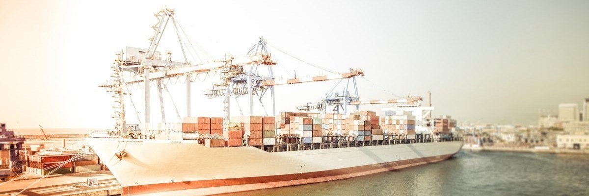 A large cargo ship loaded with colorful containers docked at a busy industrial port under bright sunlight.